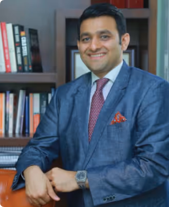 Smiling man in a blue suit with red tie and pocket square sitting with arms rested, bookshelf in background.