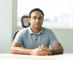 Man sitting at a desk with hands clasped, wearing a gray polo shirt, in a bright office.