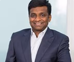Smiling man in a dark blazer and white shirt standing indoors against a light background.