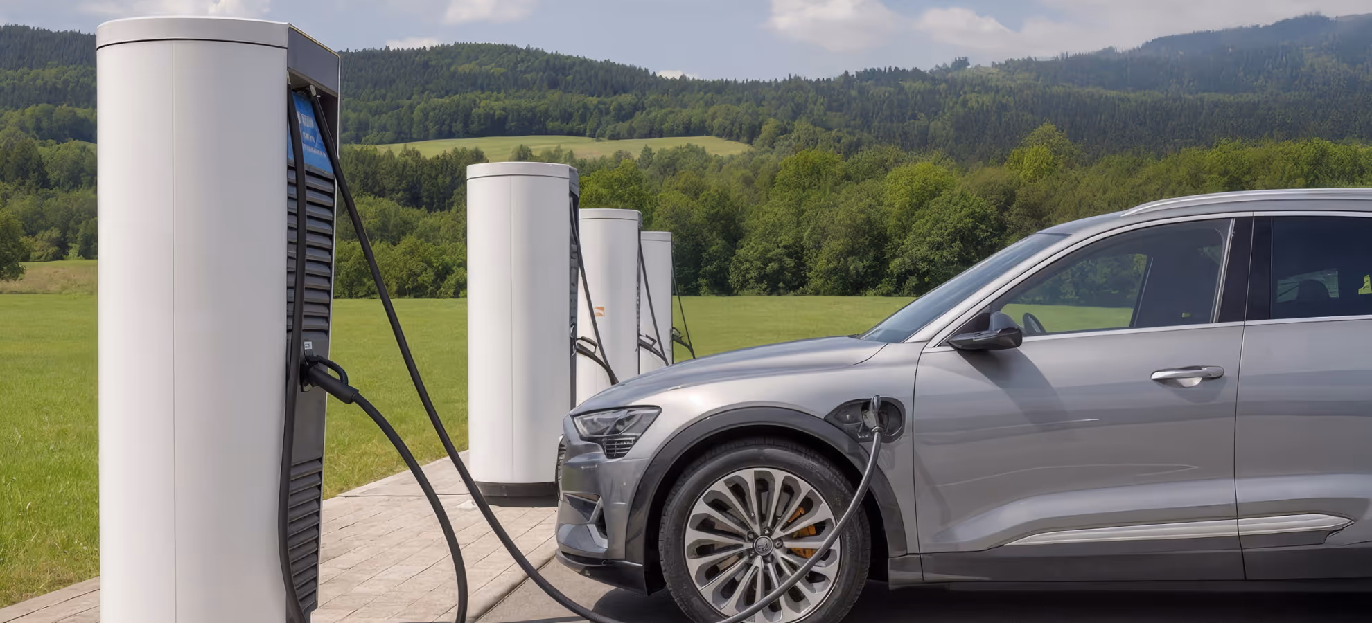 Silver electric car charging at an outdoor charging station with green trees and hills in the background.