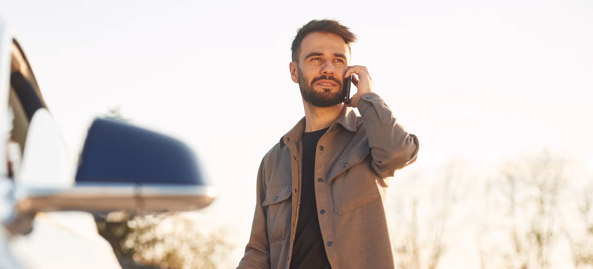 Bearded man wearing a brown jacket talking on a cellphone outdoors with a car side mirror in the foreground.
