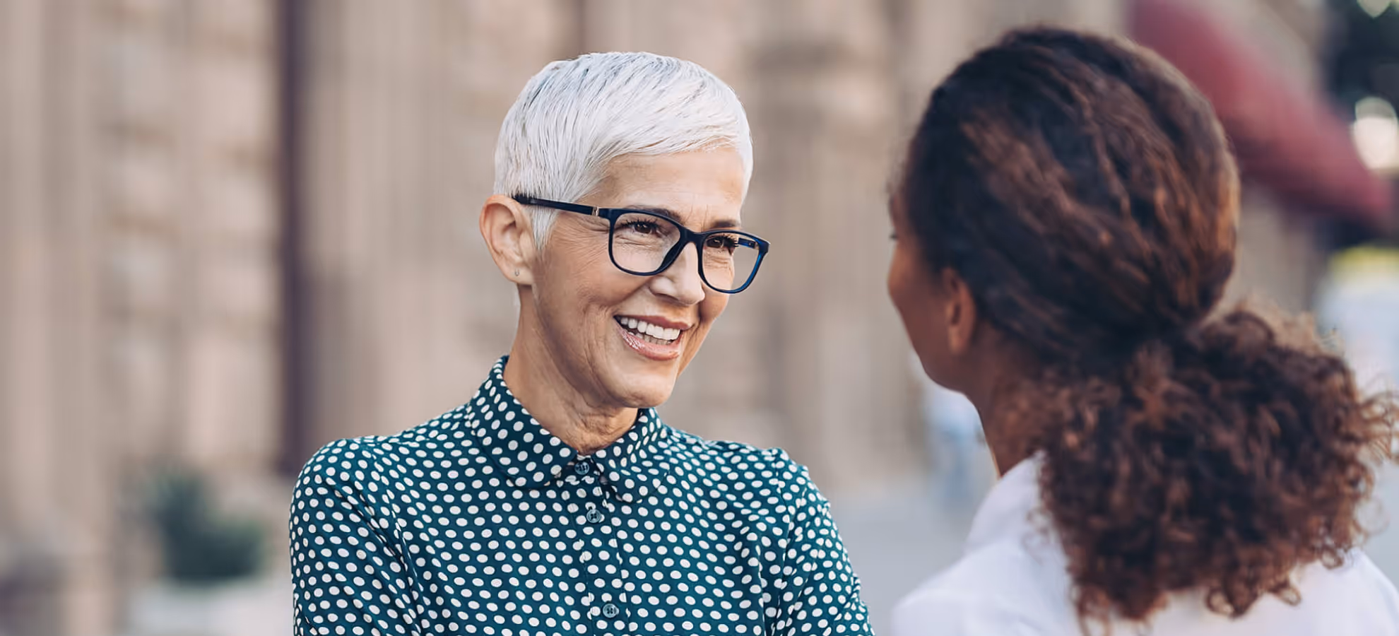 Smiling older woman with short white hair and glasses engaged in conversation with a younger woman outdoors.