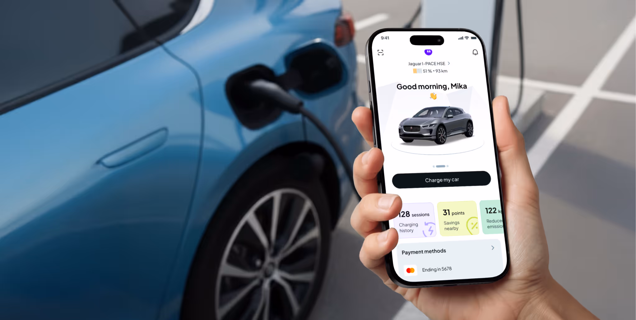 Hand holding smartphone displaying a car charging app next to a blue electric vehicle being charged.