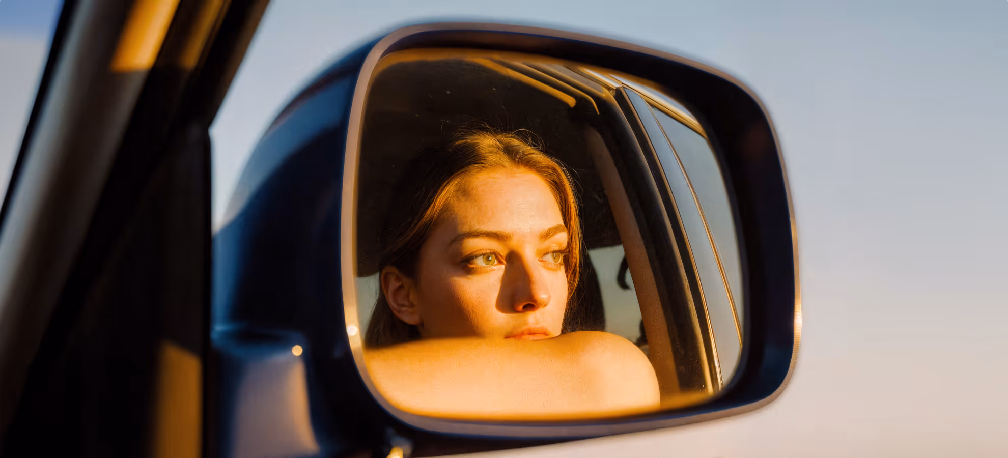 Young woman resting her chin on her arm, looking thoughtfully out of a car side mirror in warm light.