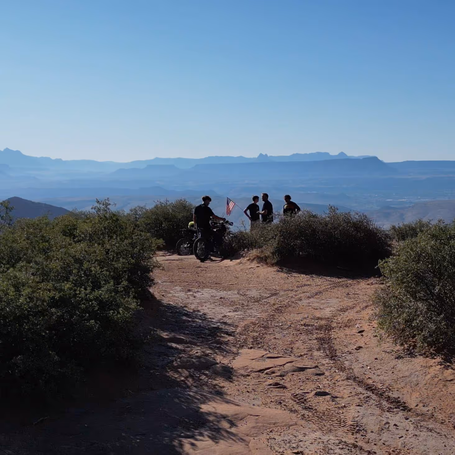 Group of four people with electric motorcycles on a rocky overlook surrounded by bushes with mountains in the background.