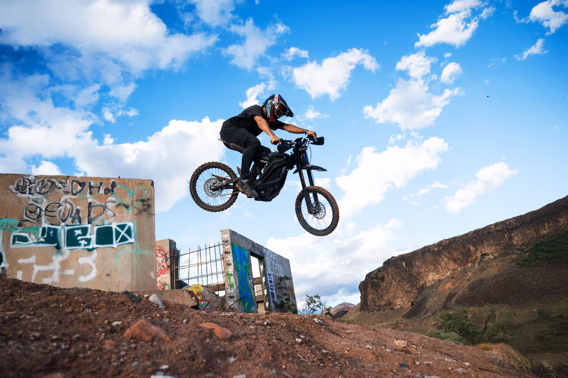 Person wearing black gear and helmet jumping an electric dirt bike over rough terrain near graffiti-covered concrete walls of the Babylon House under a blue sky with clouds.