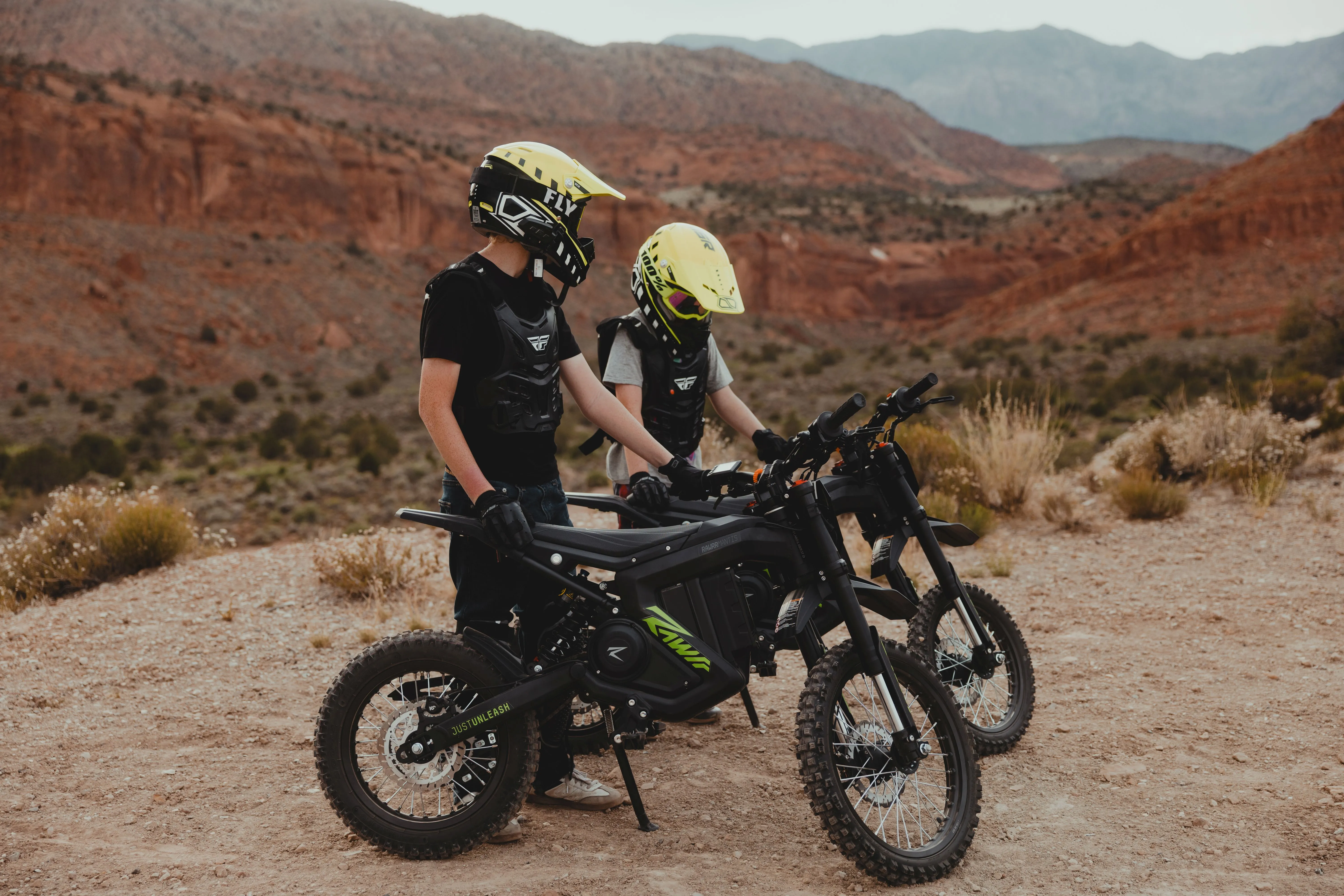 Two children wearing yellow helmets and protective gear standing with black electric dirt bikes in a desert landscape with red rocky hills.