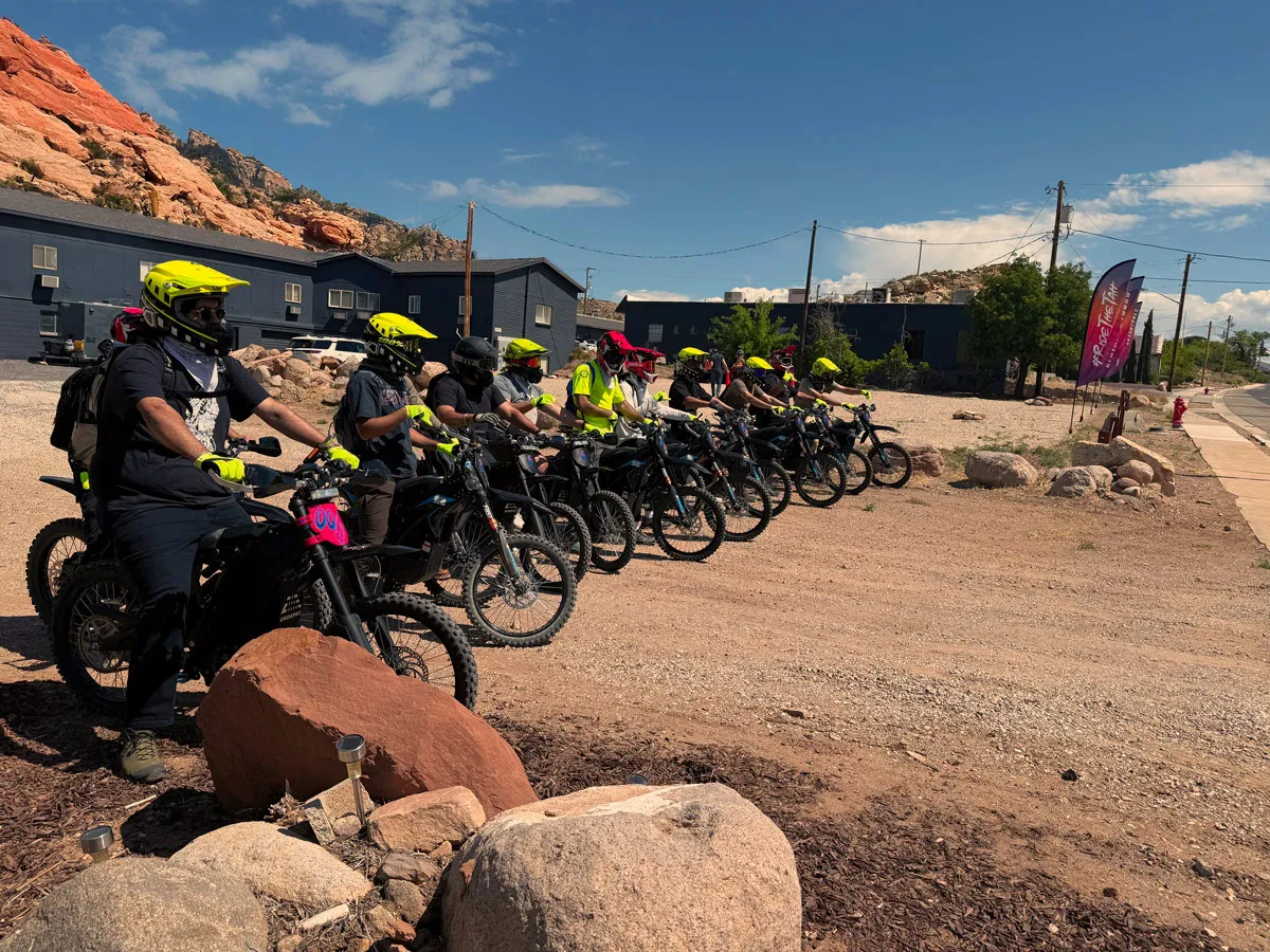 Large group of people wearing yellow and red helmets lined up on electric dirt bikes in a desert area near rocky hills and buildings.