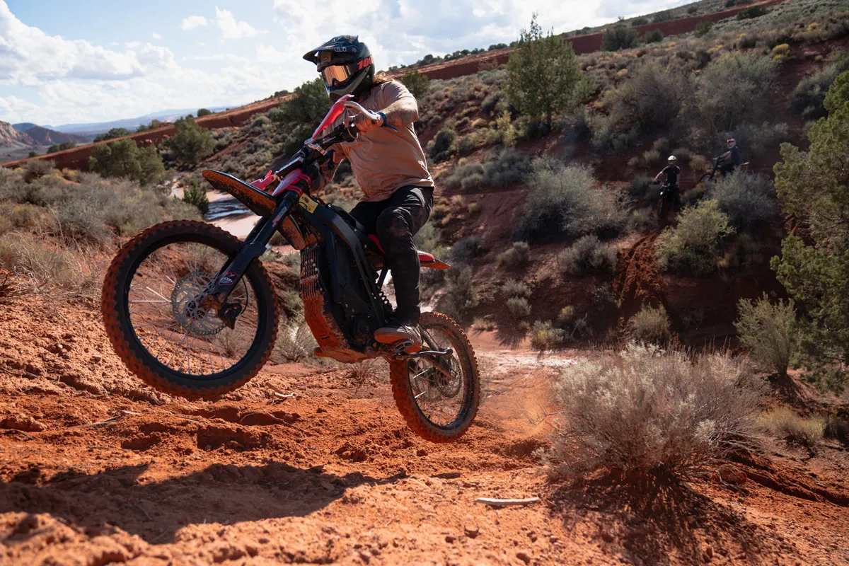 Helmeted rider riding an orange electric dirt bike rental on a red dirt desert trail.
