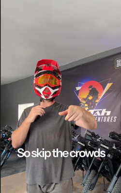 Person wearing a red and black motocross helmet and gray shirt pointing towards the camera in a bike shop with bicycles and an 'Utah Adventures' sign in the background.