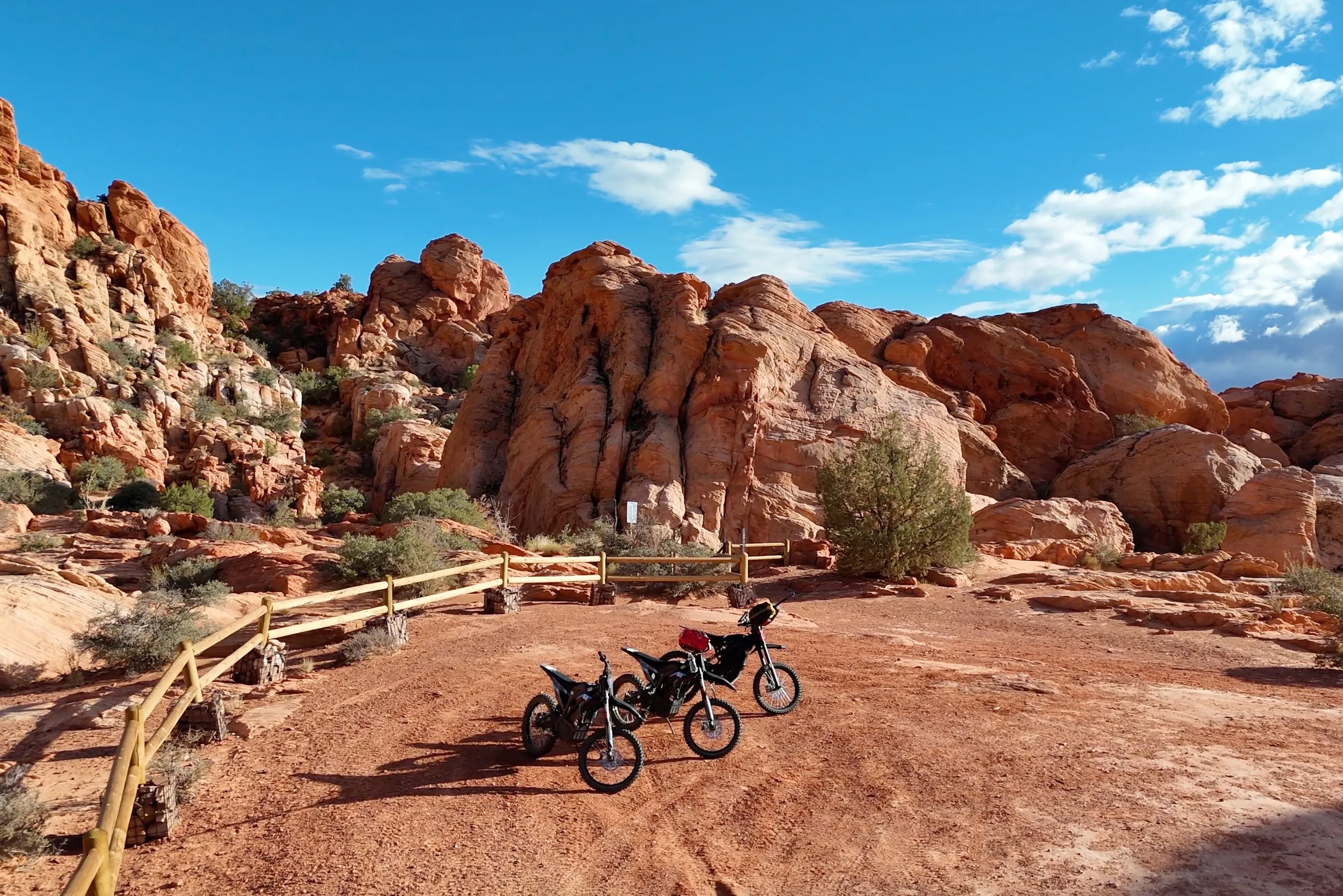 Sunny desert landscape with red rock formations, wooden fence, and three parked dirt bikes.