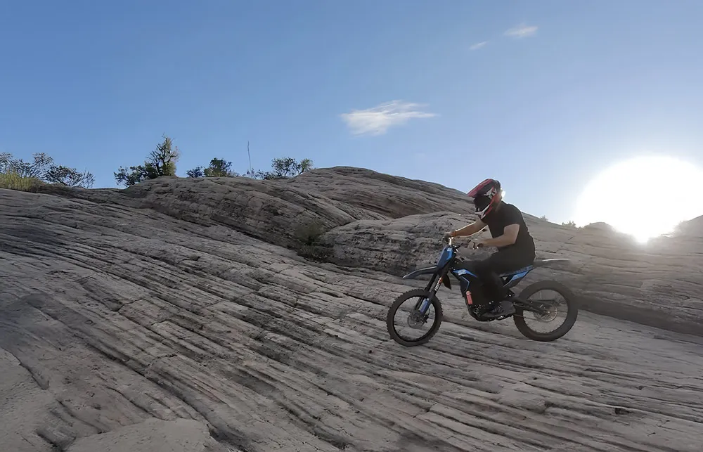 Person wearing helmet riding a dirt bike on rocky terrain under a clear blue sky with sun low on horizon.