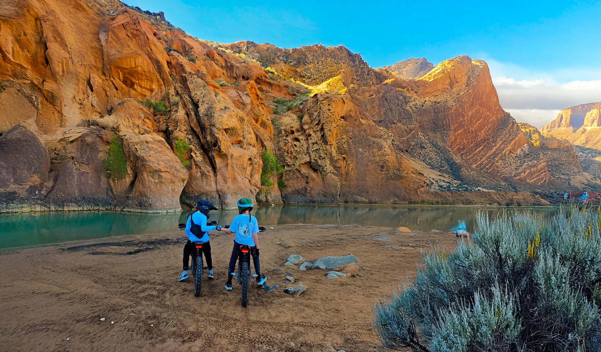 Two kids on dirt bikes fist-bumping near a river with rocky cliffs and blue sky in the background.