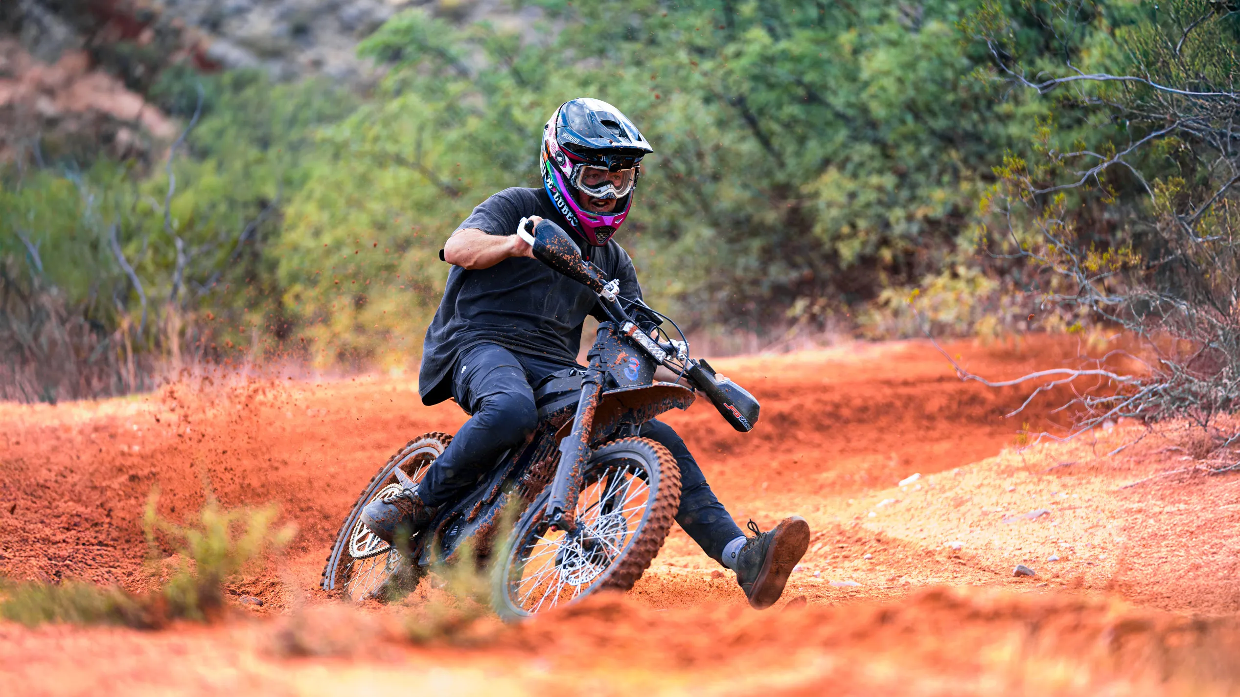 Motorcyclist in black gear riding a dirt bike kicking up orange dirt on a trail surrounded by green bushes.