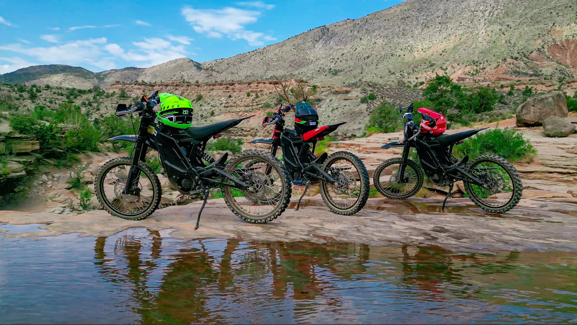 Three dirt bikes with helmets parked on rocky terrain with mountains and blue sky in background.