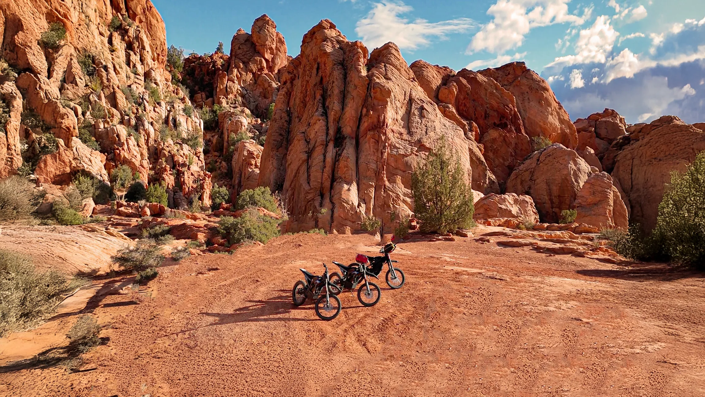Two electric dirt bikes parked on reddish dirt terrain with large rocky cliffs and blue sky background.
