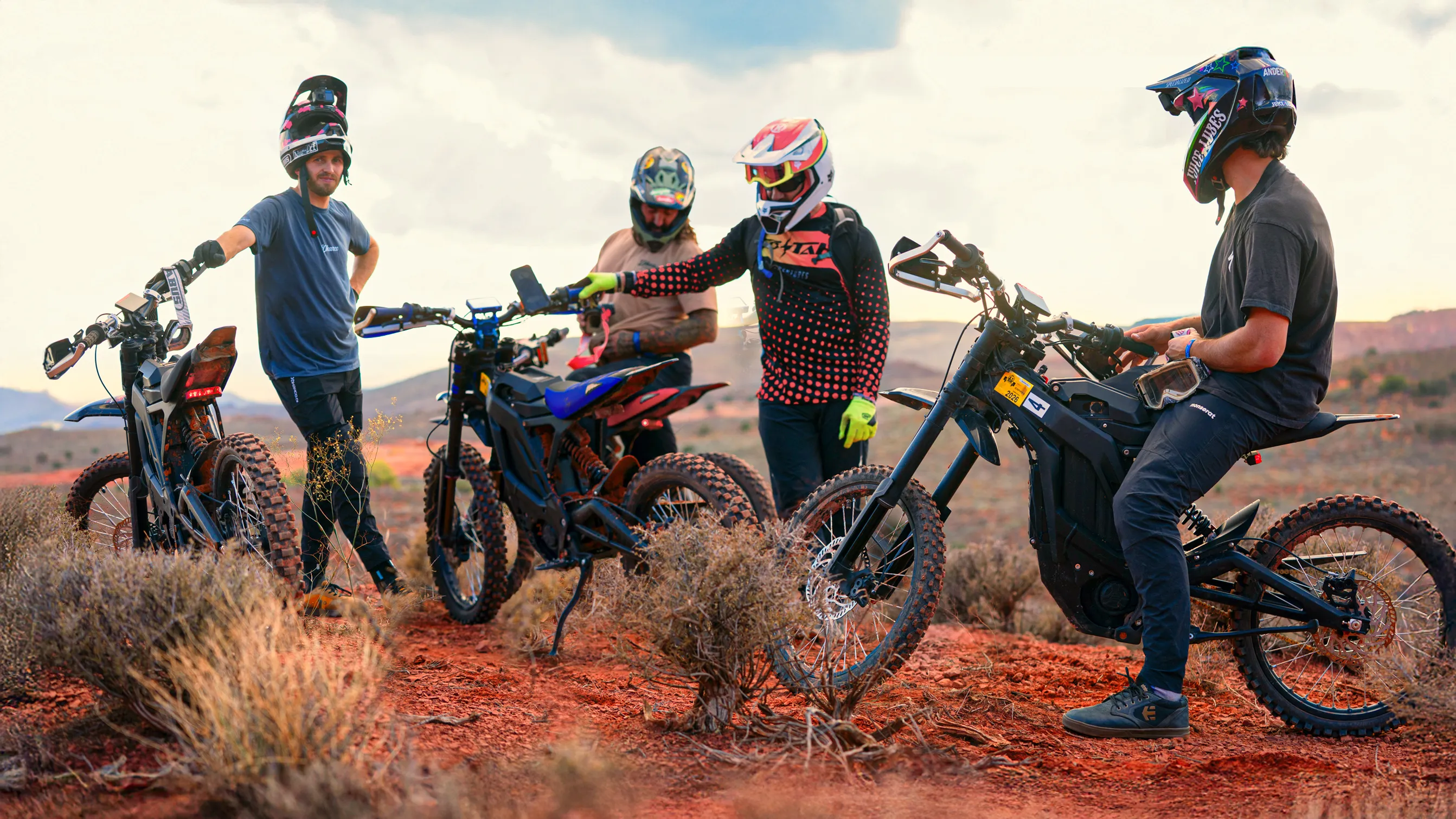 Four dirt bikers in helmets with bikes on a dry, red dirt trail in a desert landscape.