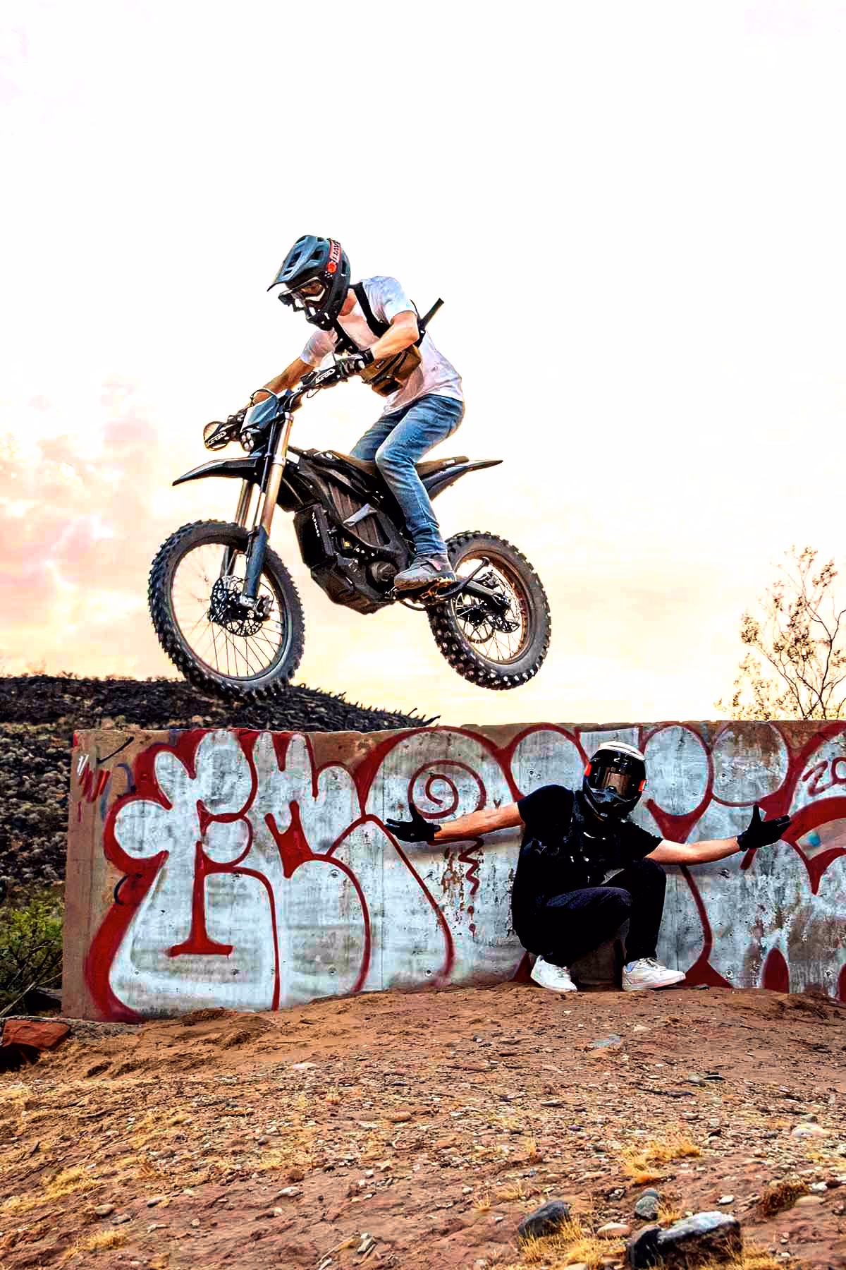 Motorcyclist wearing helmet jumps over a graffiti-covered wall while another person crouches nearby.