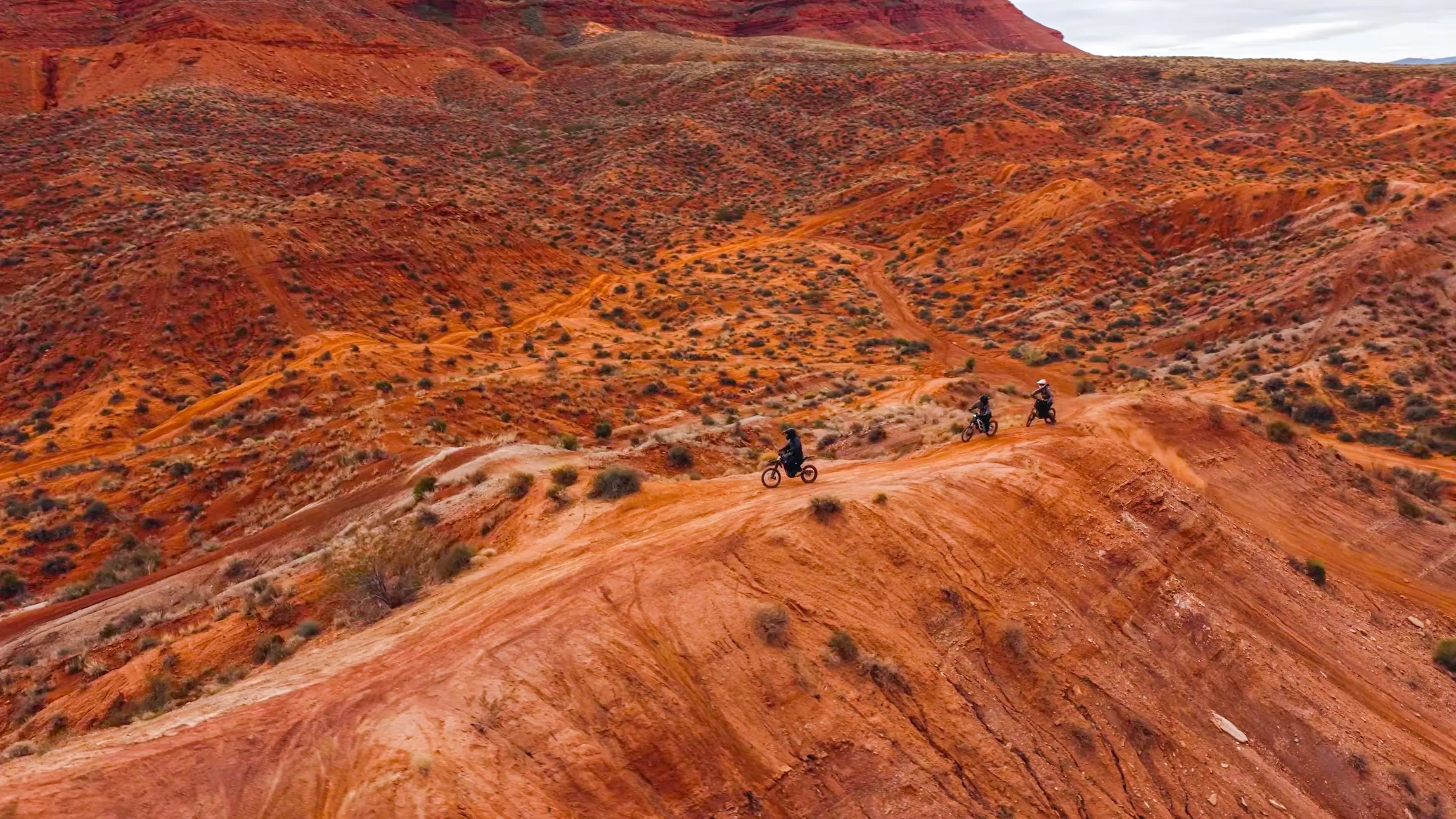 Aerial view of two riders on electric dirt bikes following a narrow ridge trail through red rock hills near Hurricane, Utah