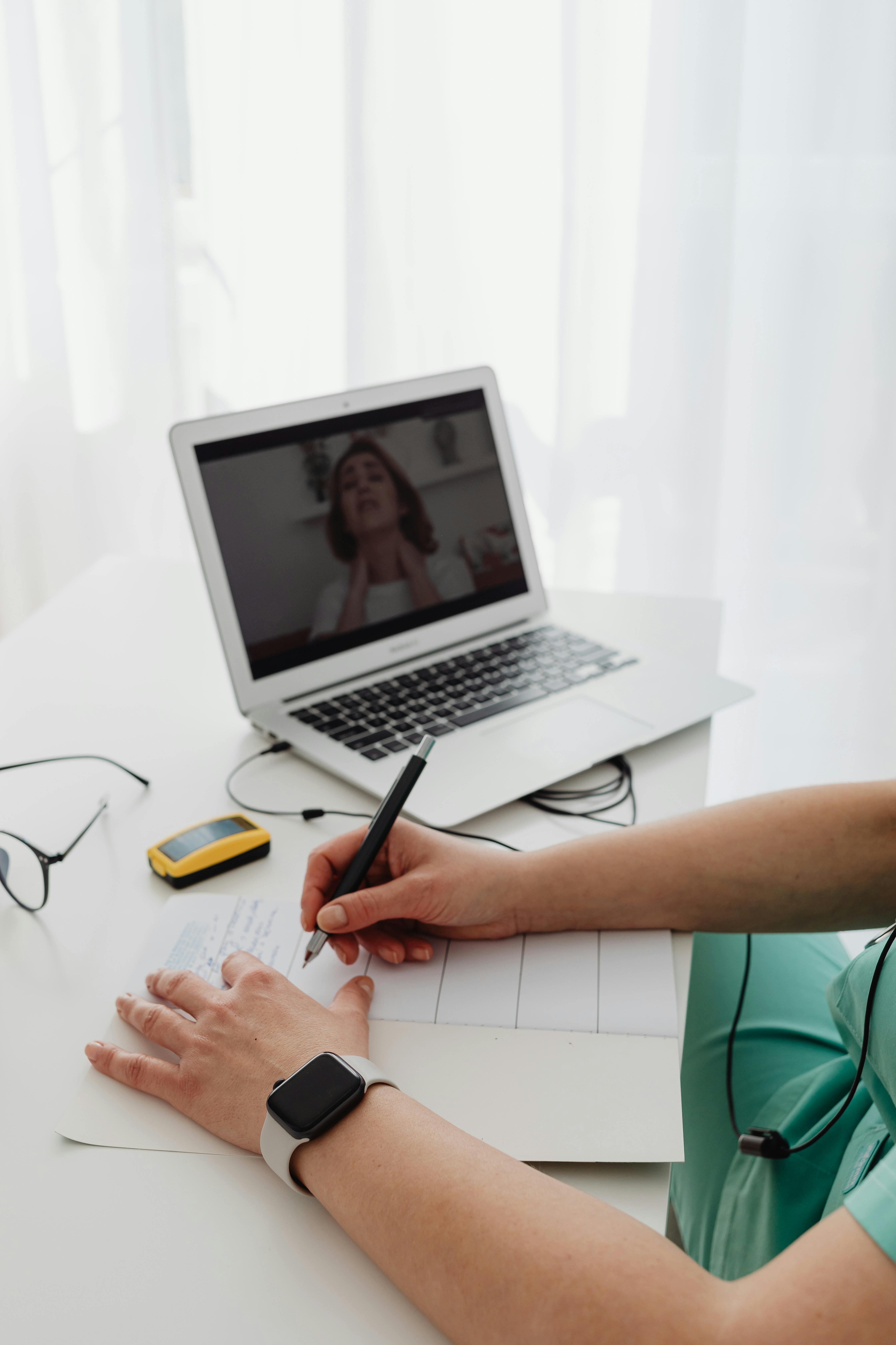 Person wearing a smartwatch taking notes in a notebook with a laptop showing a video call in the background.
