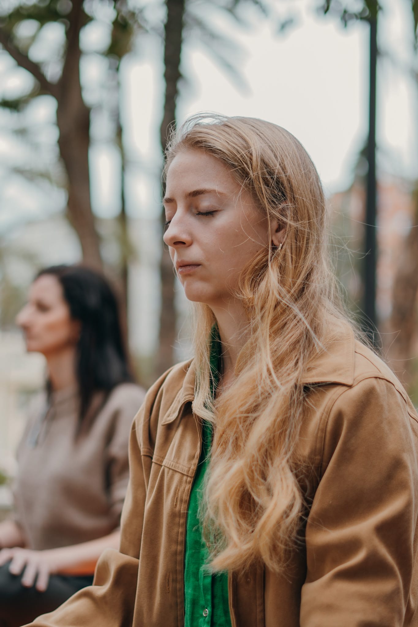 Two women sitting outdoors with eyes closed, meditating in a natural setting with blurred trees in the background.