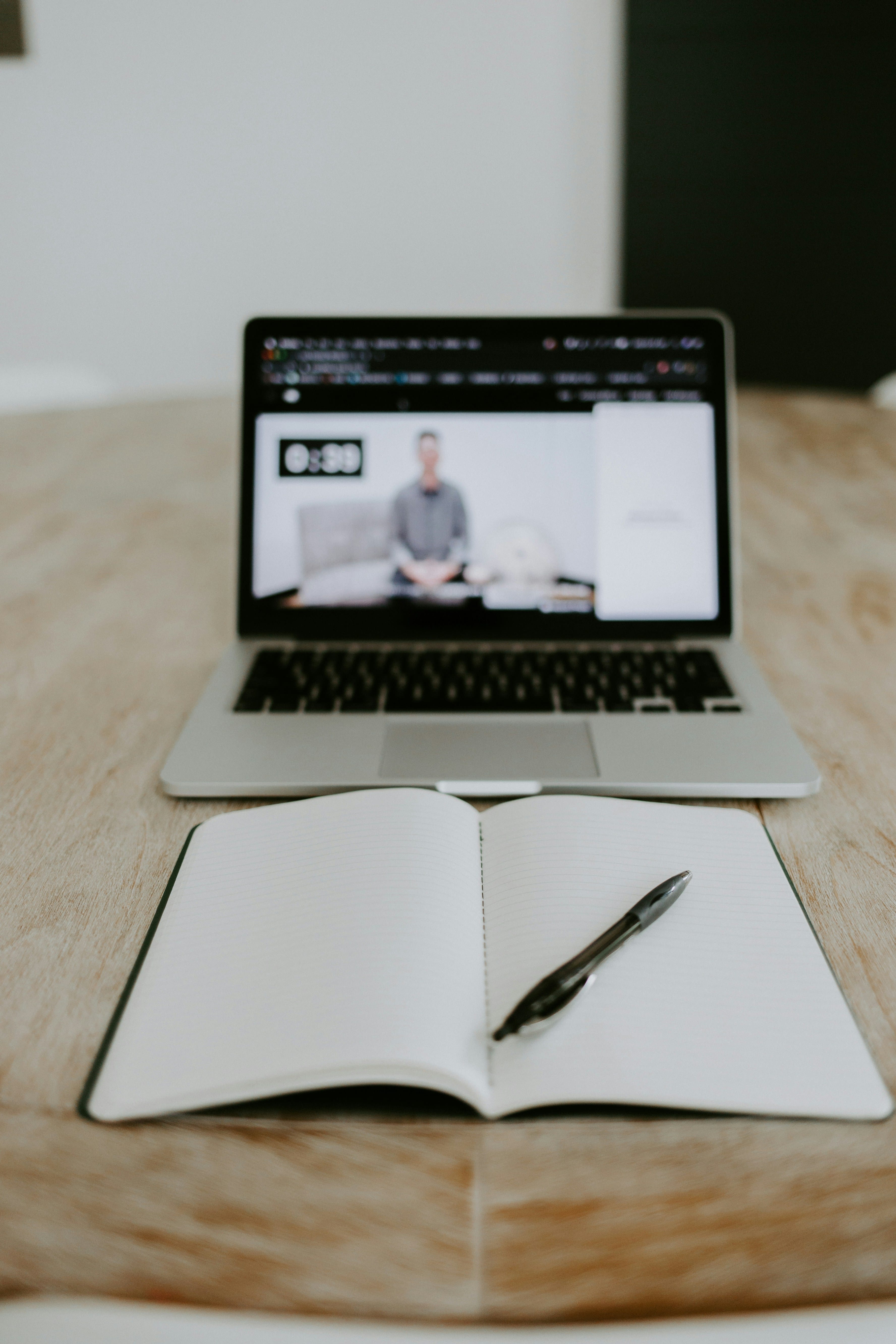 Open notebook with a black pen on a wooden table in front of a laptop playing a video of a person sitting on a sofa.
