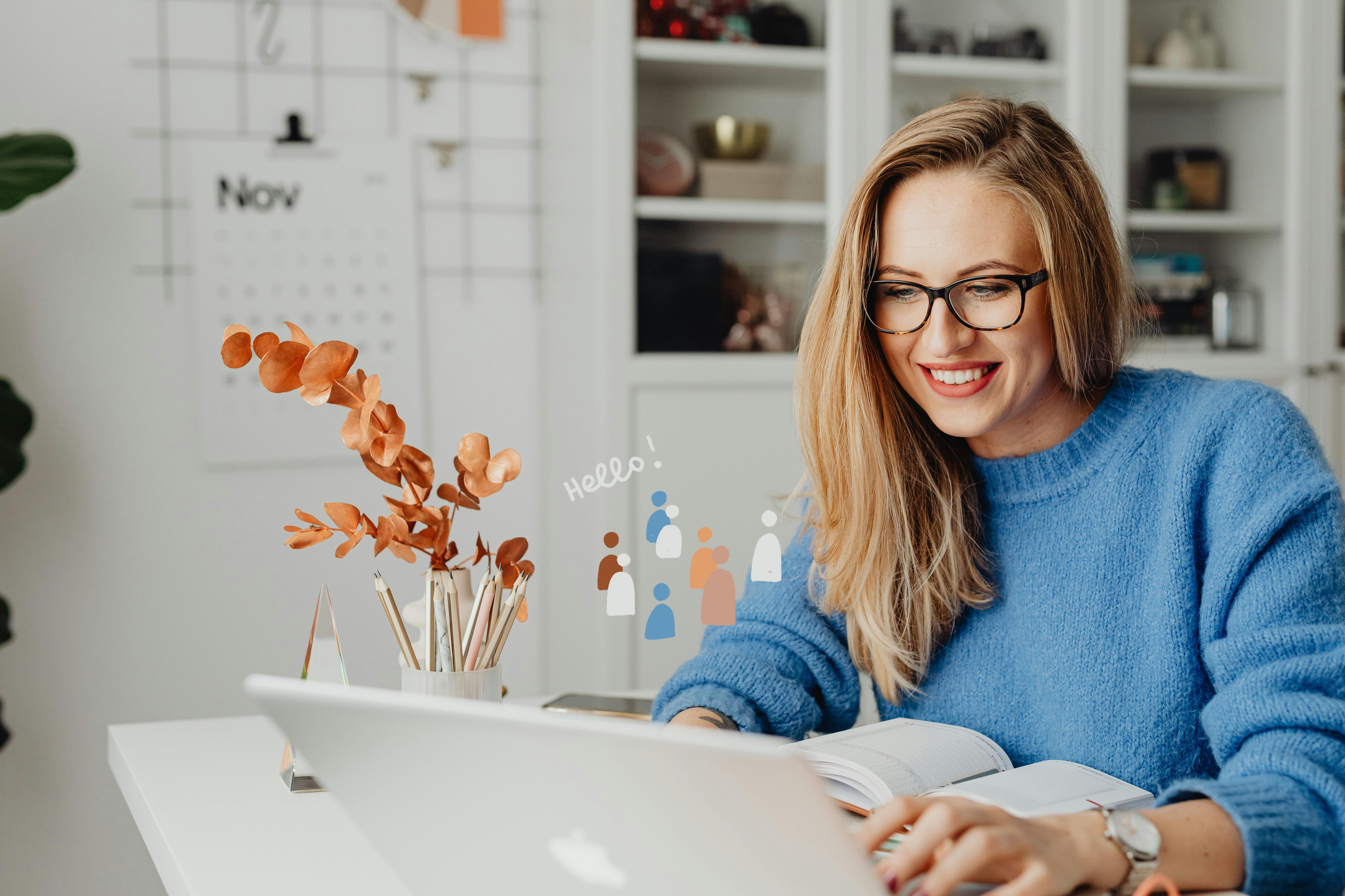 Smiling woman with glasses in a blue sweater working on a laptop at a white desk with an open notebook and pencil holder.