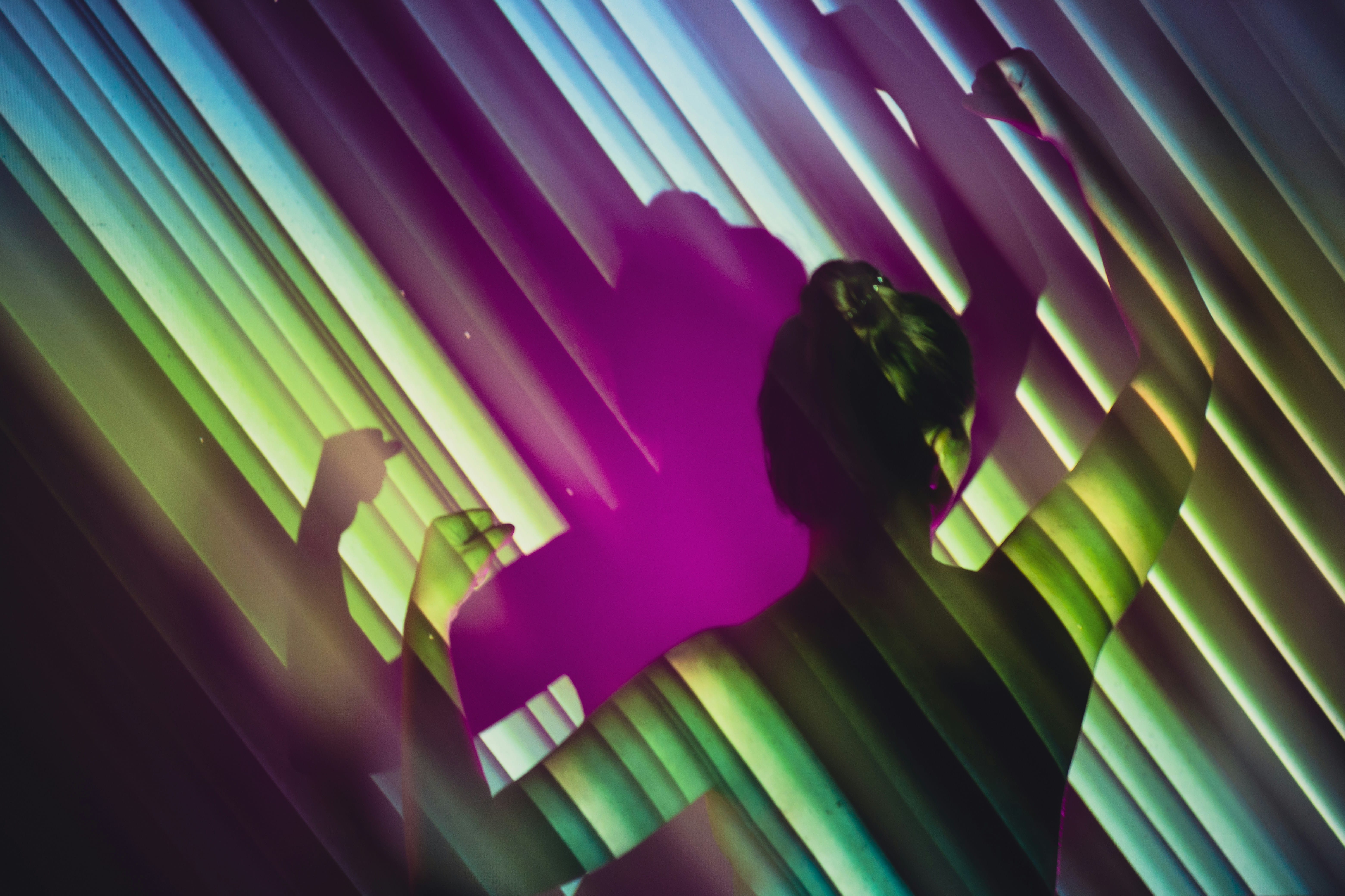 Silhouette of a person with raised arms projected with colorful striped light patterns in purple, green, and blue tones.