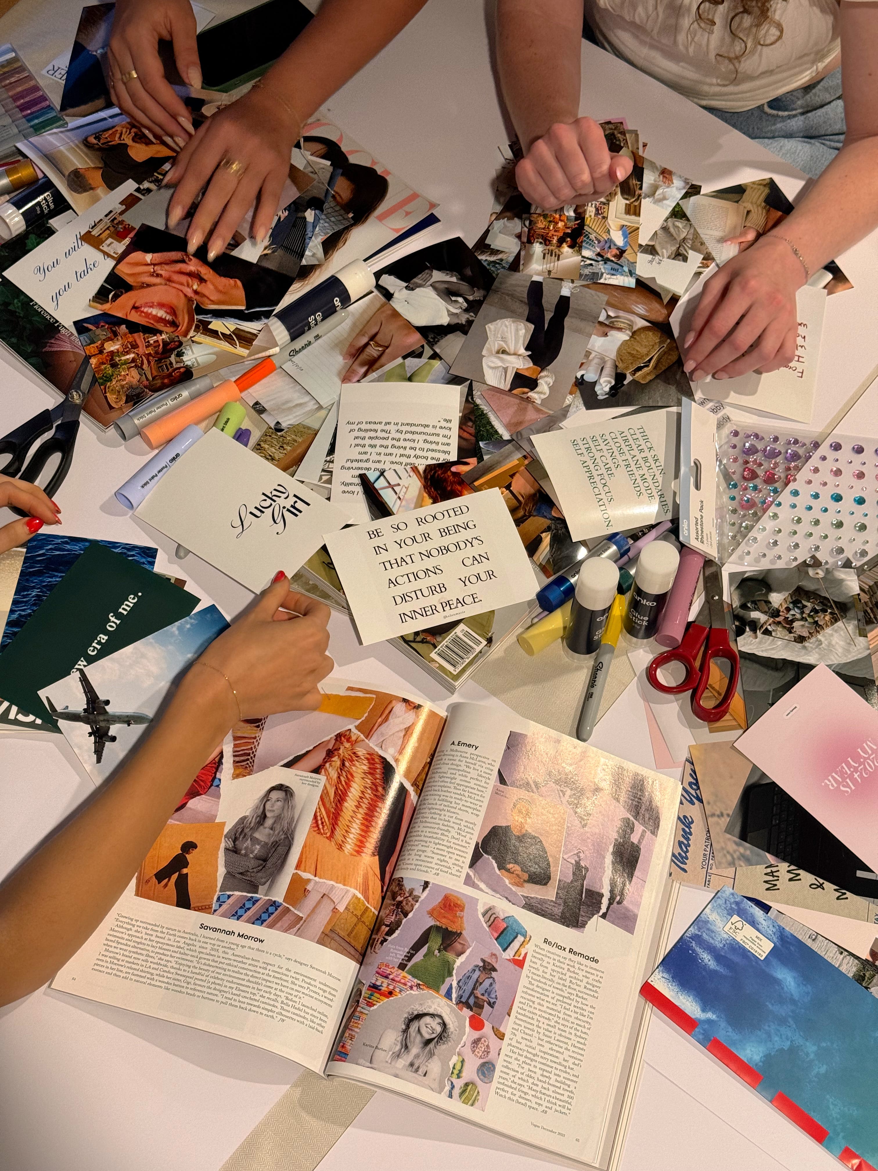 Four hands working on a collage with magazine cutouts, photos, quotes, markers, glue, scissors, and colorful craft supplies spread on a white table.
