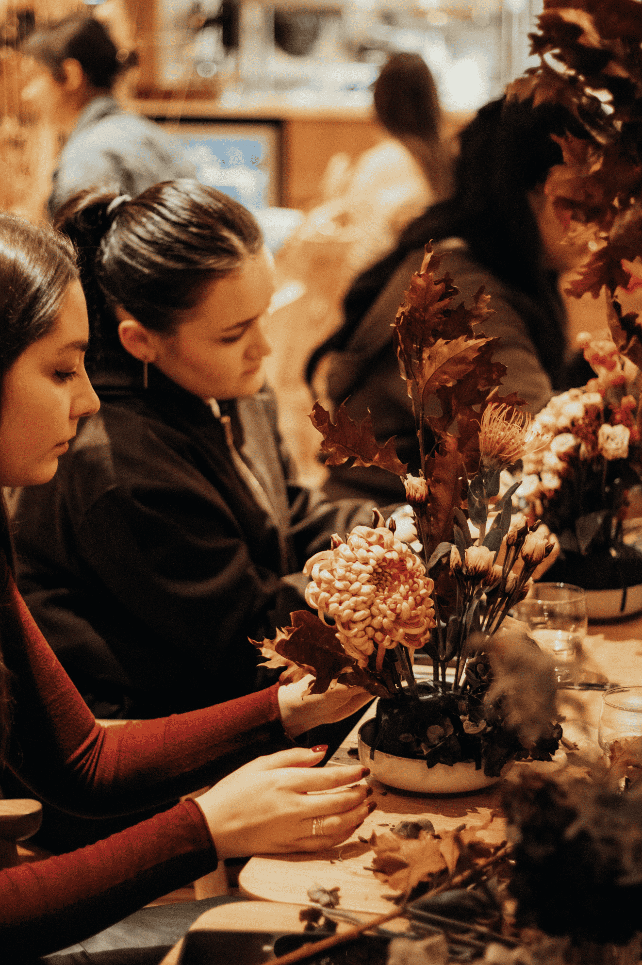 Woman with curly hair sitting cross-legged on a yoga mat with eyes closed, surrounded by other people in a calm indoor setting.