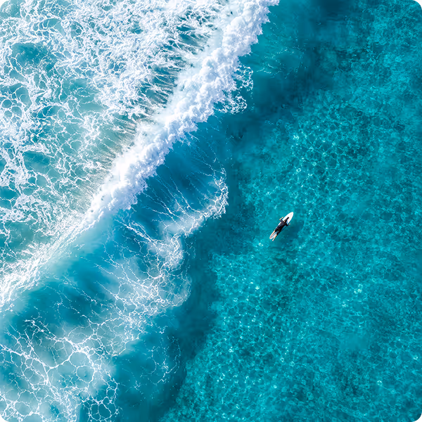 Aerial view of a surfer paddling on a board in turquoise ocean near white waves.