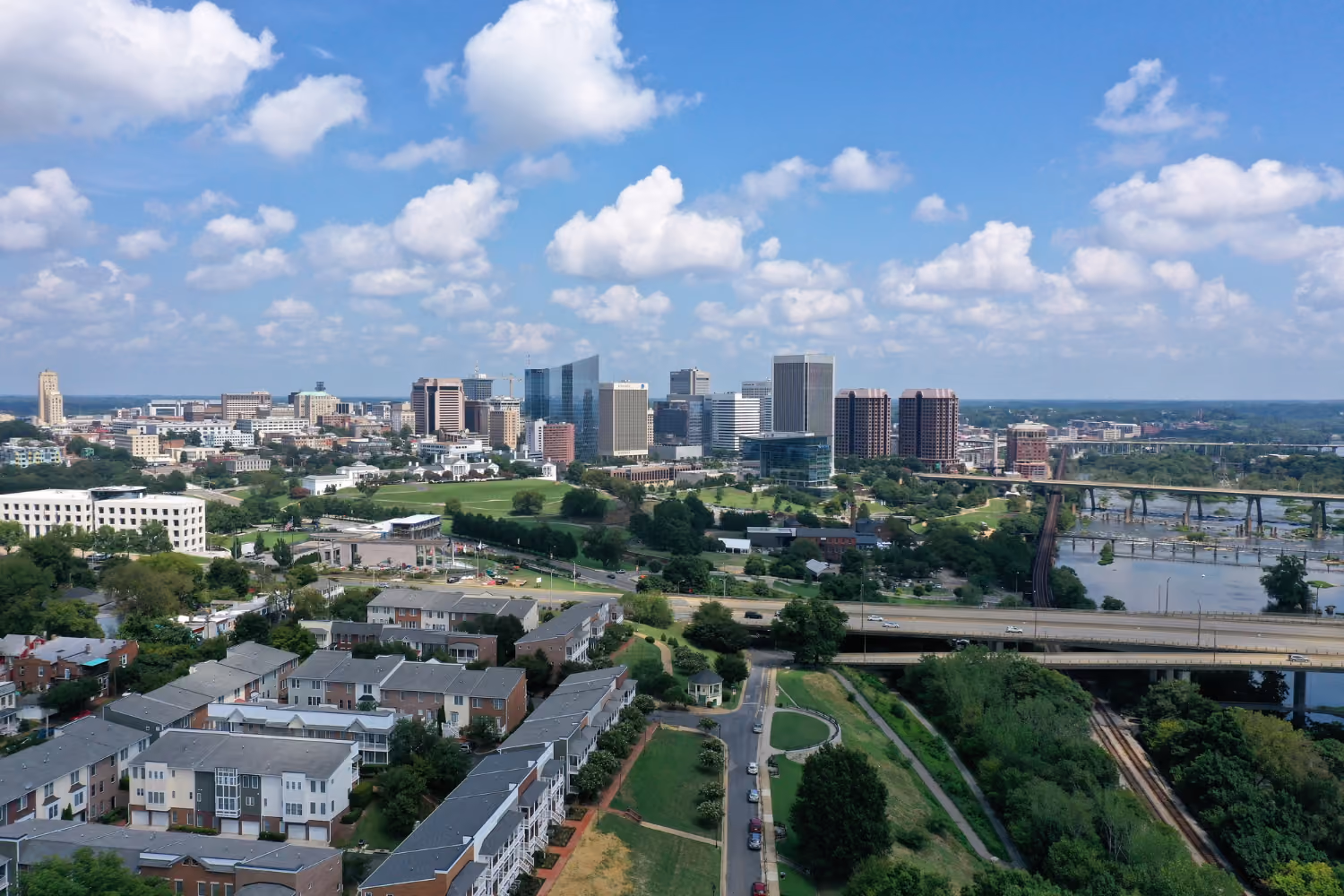 A cityscape under a blue sky with scattered clouds, featuring a cluster of mid- to high-rise buildings in the distance, residential houses in the foreground, green spaces, roads with some cars, and bridges crossing a river on the right.