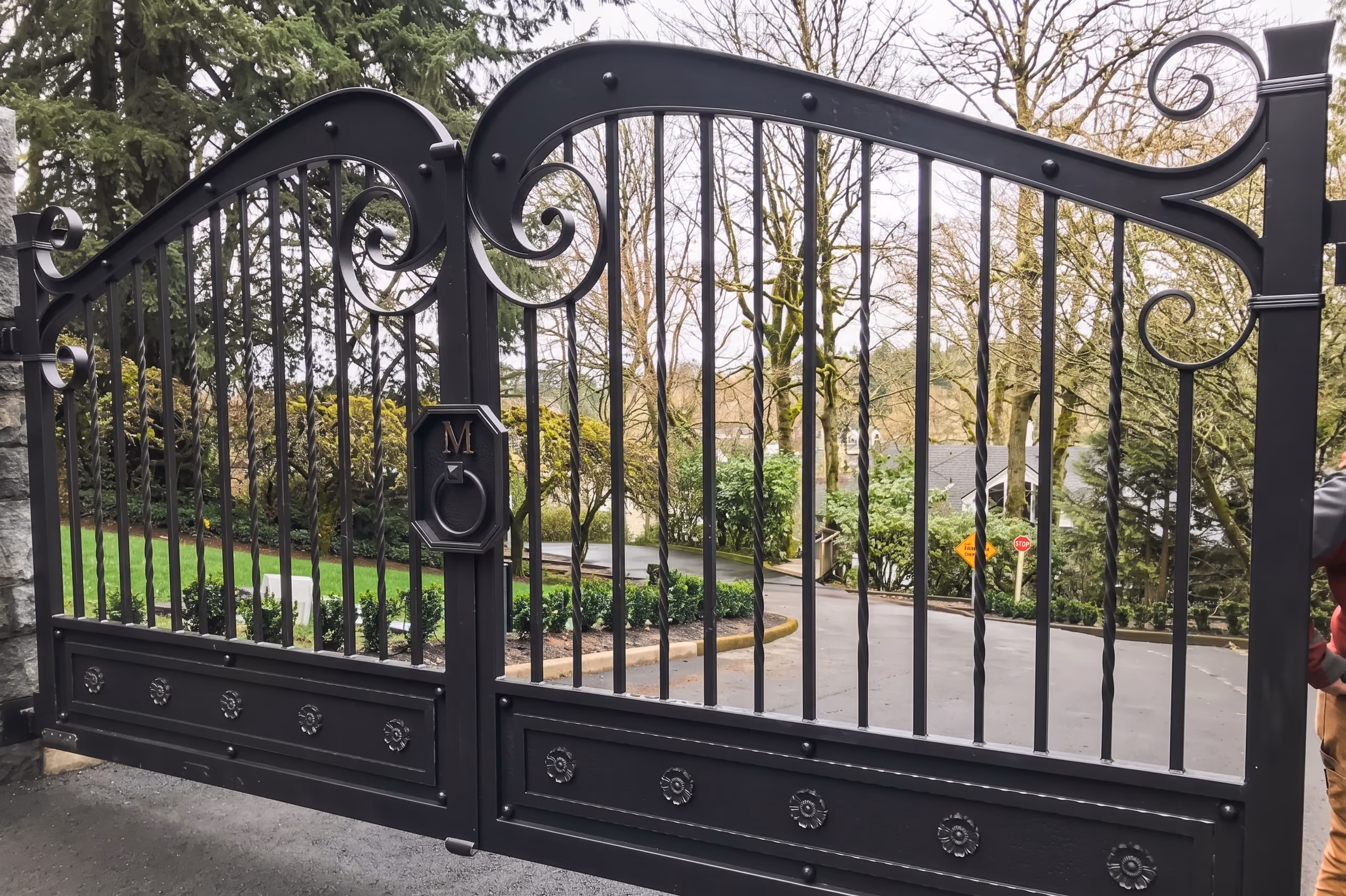 Closed black wrought iron gate with vertical bars, decorative scrolls, and floral patterns at the bottom panels, set at the entrance to a residential area with trees and greenery visible behind.