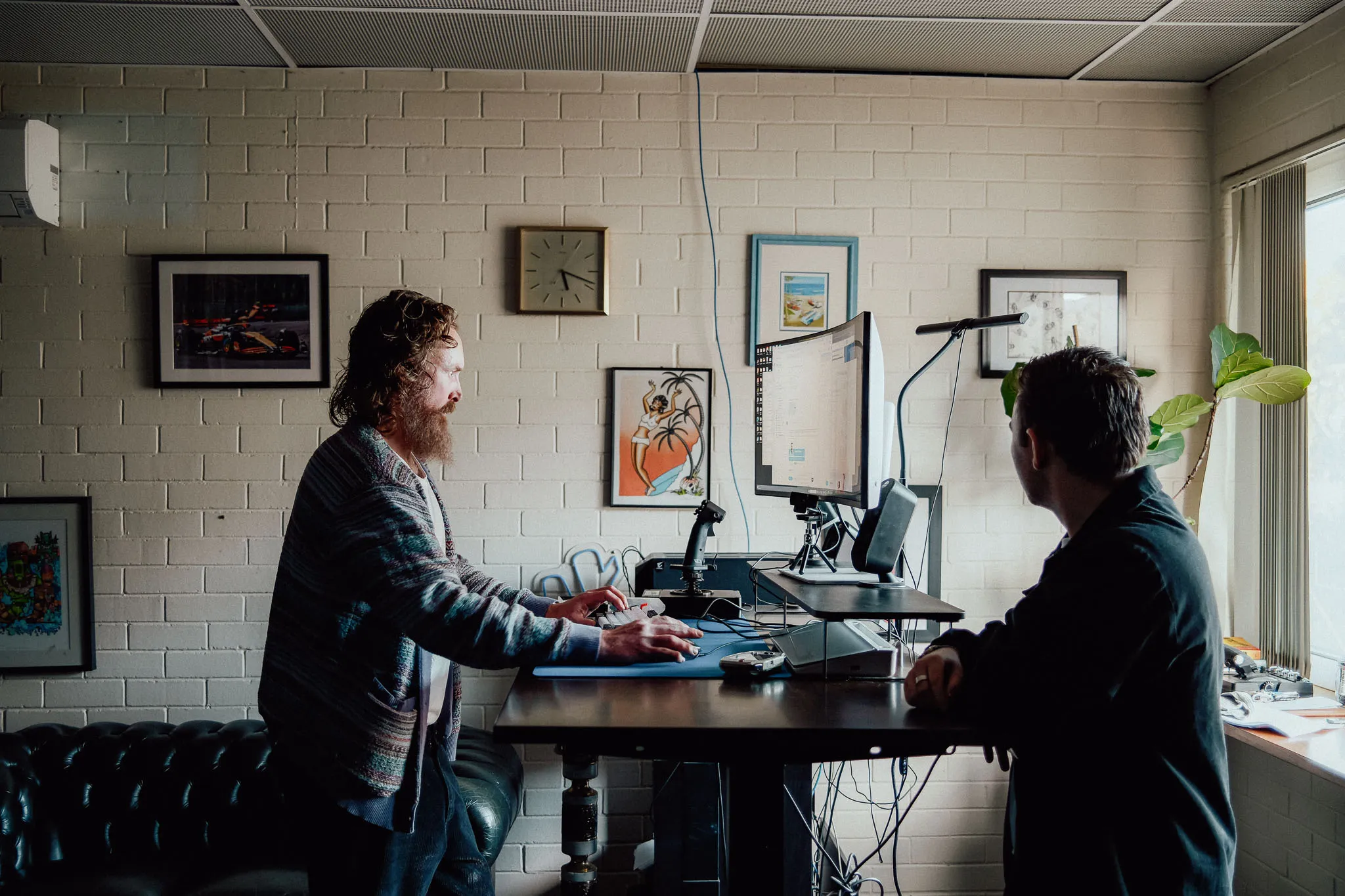 Two men in an office working at a standing desk with a computer monitor and framed pictures on the wall.