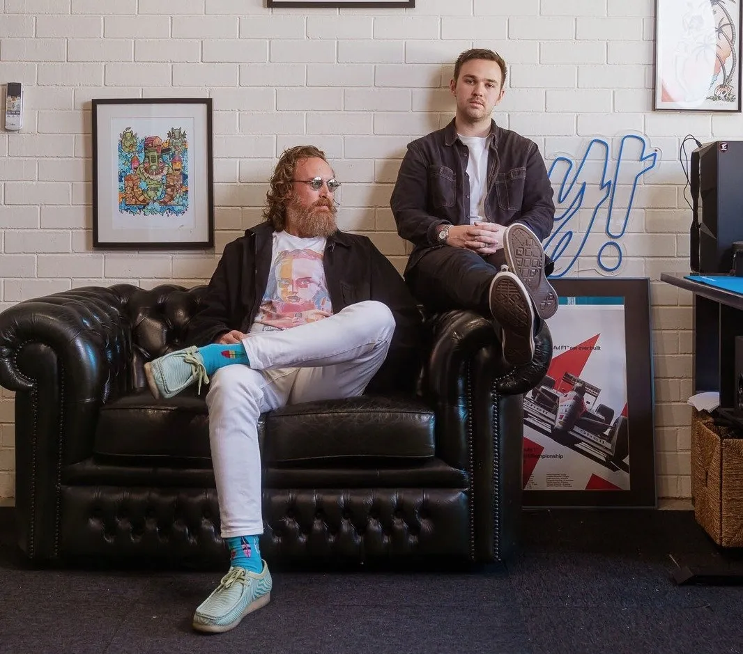 Two men sitting on a black leather couch against a white brick wall with framed art behind them.