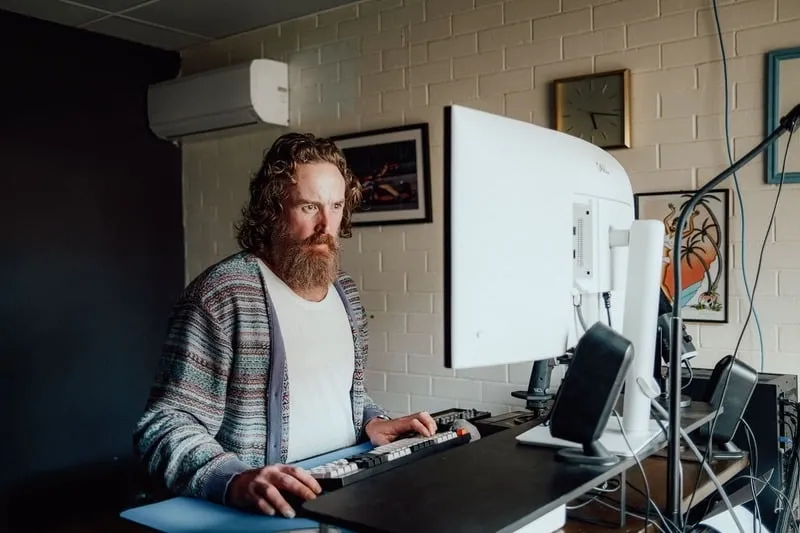 Bearded man wearing a striped cardigan working at a desk with a large monitor and keyboard.