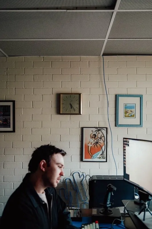 Man sitting at desk with computer and joystick, beige brick wall with clock and framed pictures behind him.