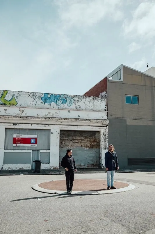 Two men standing apart on a circular sidewalk area in front of a weathered building with graffiti.