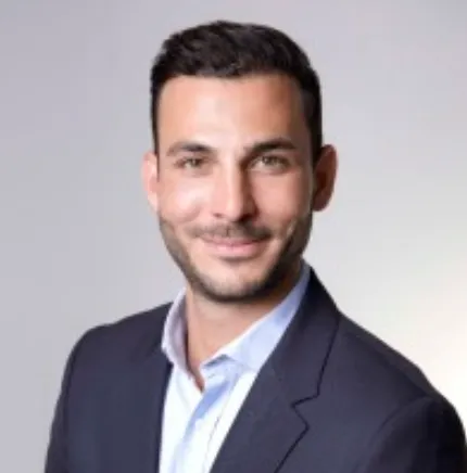 Smiling man with short black hair wearing a dark blazer and light blue shirt against a gray background.