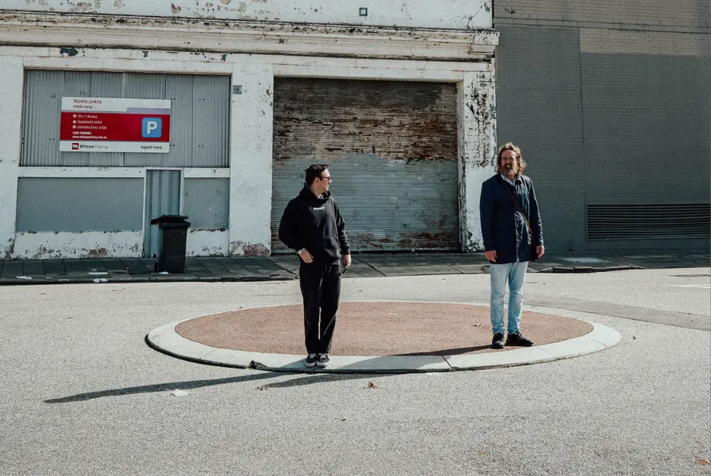 Two men standing apart on a circular patch in an empty urban parking area with a weathered building behind.