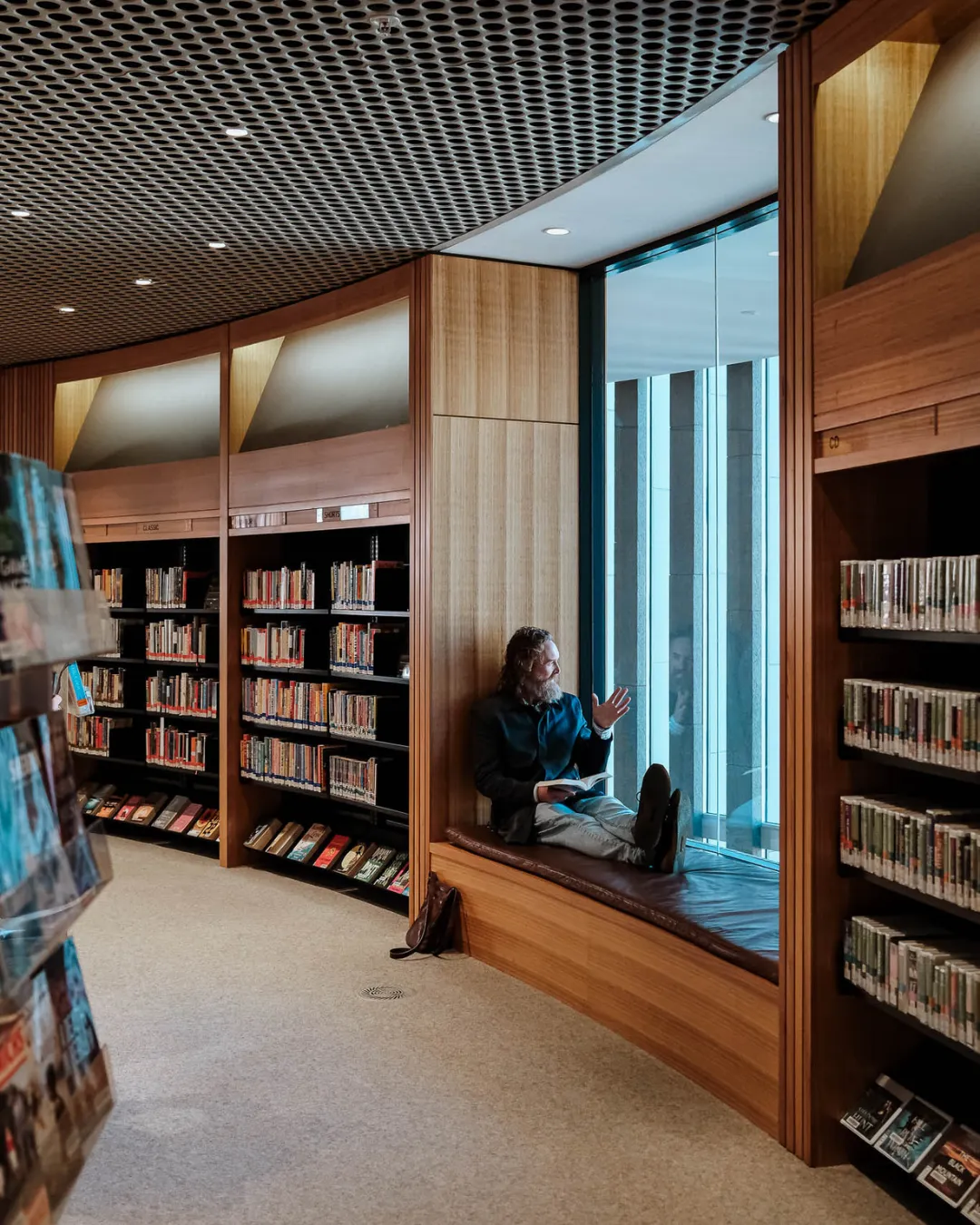 Man sitting on a window seat reading a book in a modern library with wooden bookshelves.