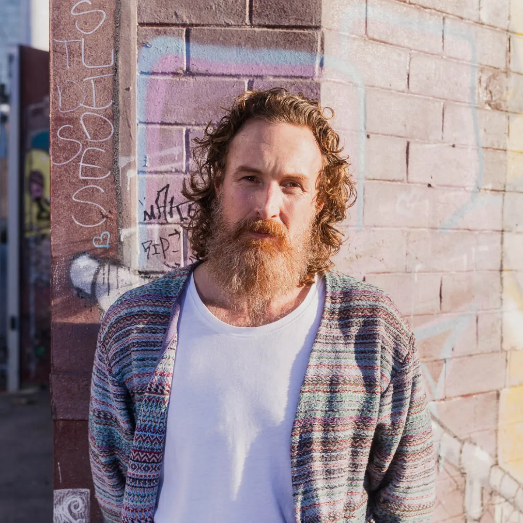 Man with curly hair and long beard wearing a patterned cardigan and white shirt by a graffiti wall.