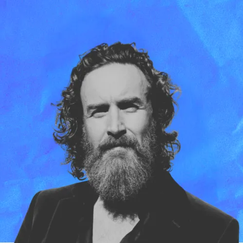 Portrait of a bearded man with curly hair in a black jacket against a blue background.