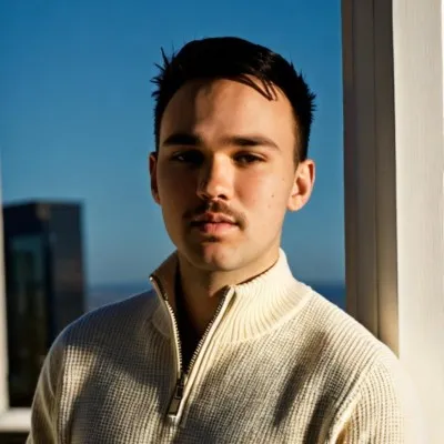 Young man with short hair and mustache wearing a white sweater, standing by a window with city view.