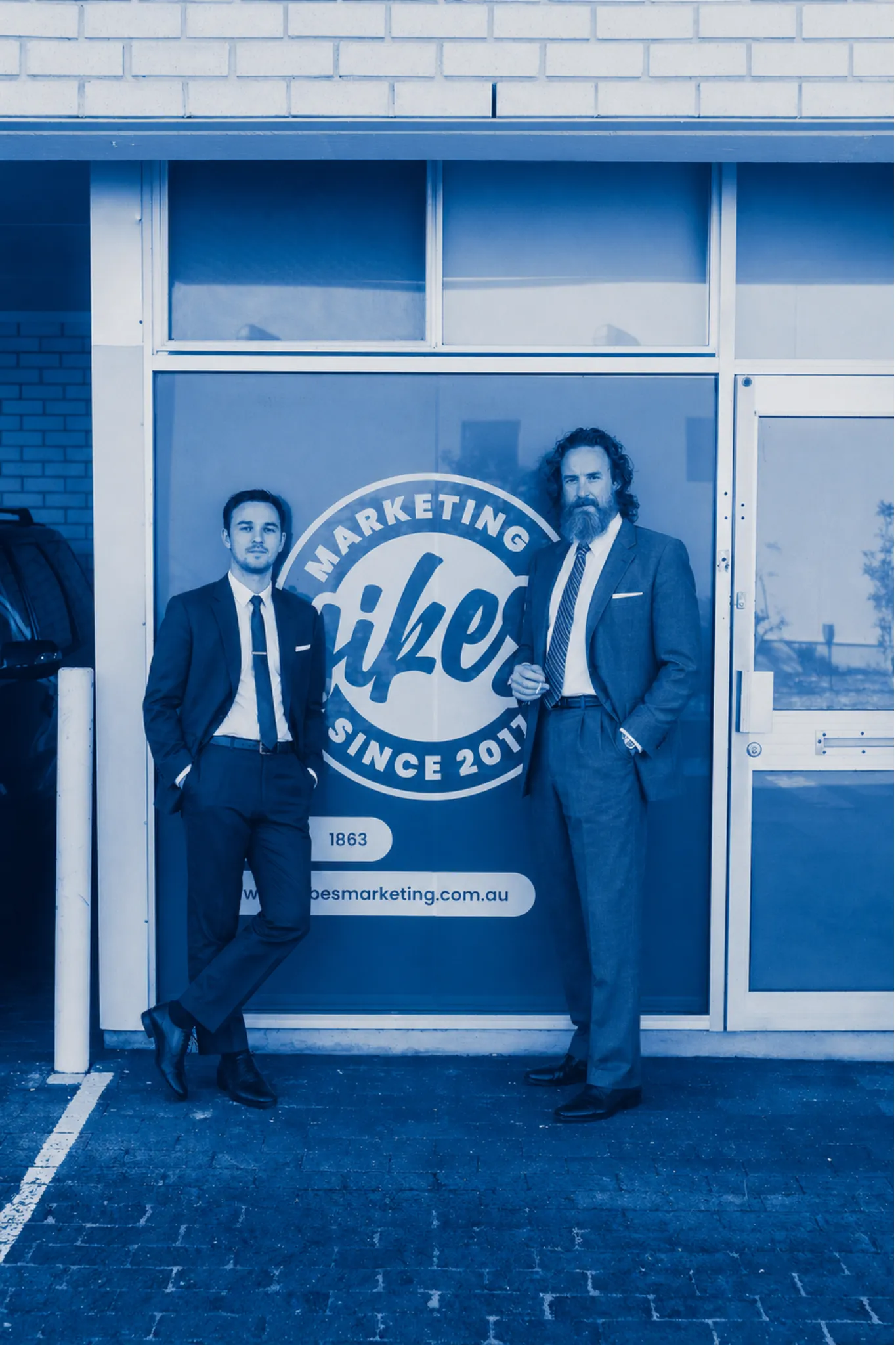 Two men in suits standing outside a marketing office with a large window sign.