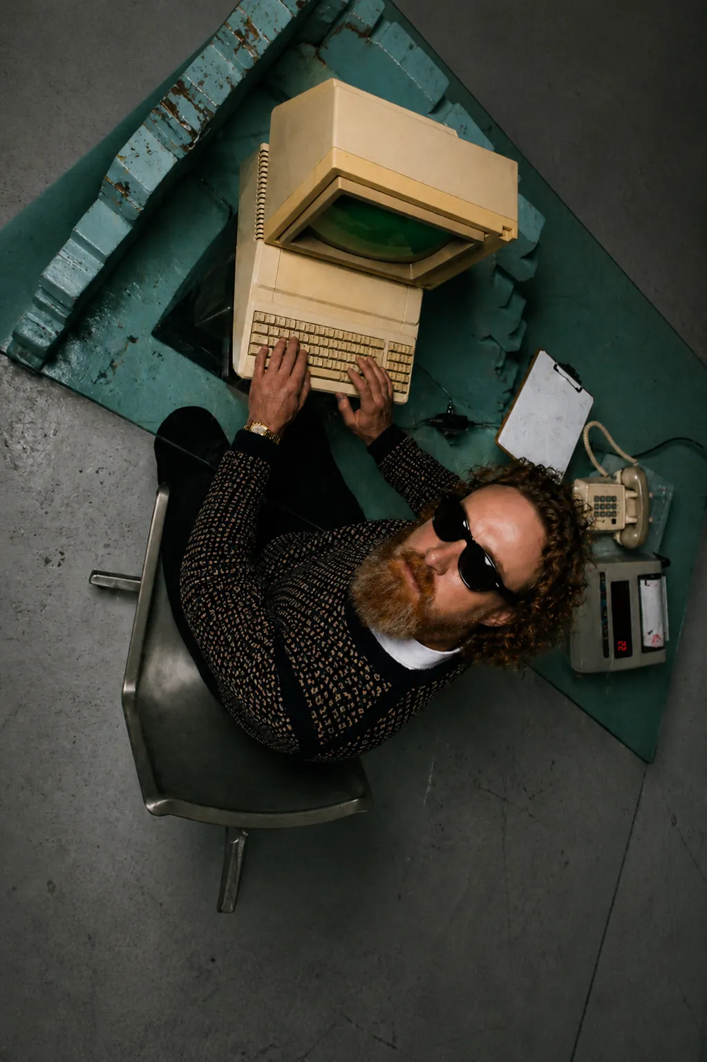 Man with curly hair and sunglasses typing on a vintage computer at a blue desk with a phone and calculator.