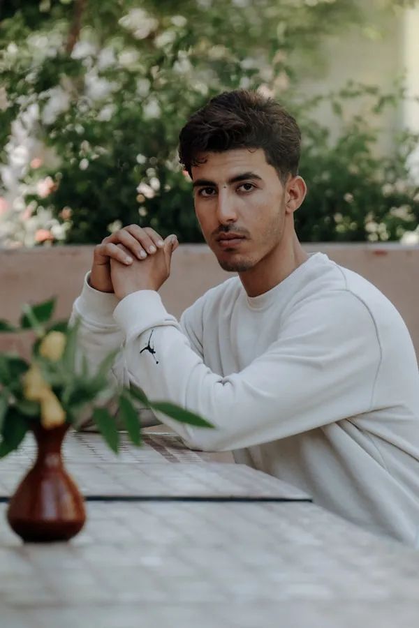 Young man in a white sweatshirt sitting at a tiled table outdoors with hands clasped.