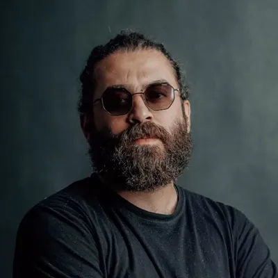Man with beard and sunglasses wearing a black shirt against a dark plain background.