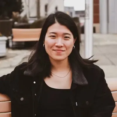 Young woman with long black hair wearing a black jacket, sitting on a bench outdoors, smiling.