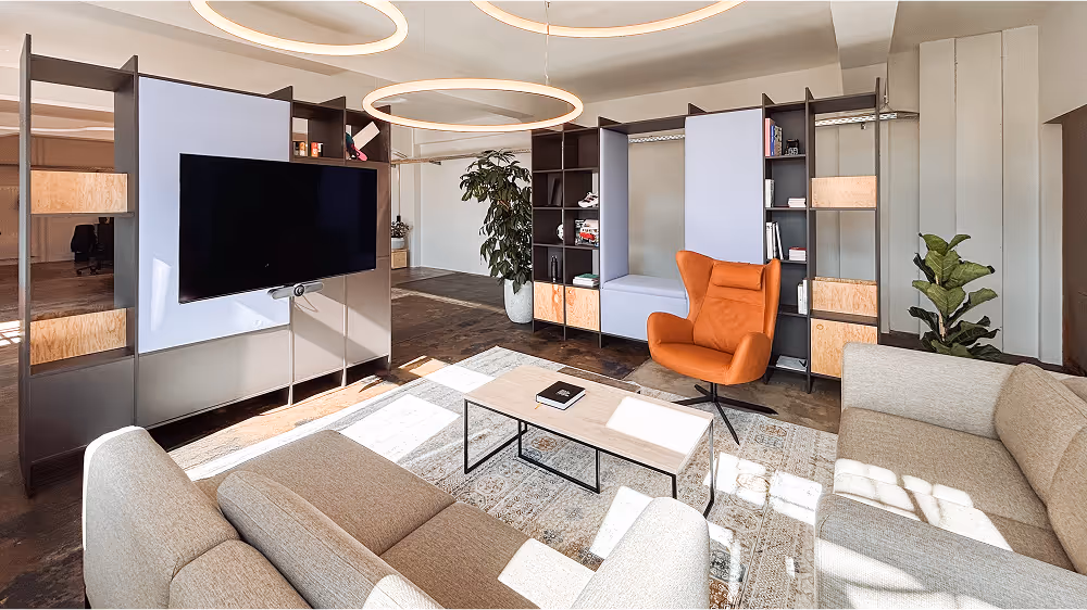 Our office lounge in Munich with light-colored sofas, an orange armchair, a coffee table, and a large TV on a shelf. Room dividers with books and plants separate the lounge area from the workspace.